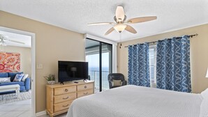 Une chambre à coucher avec un lit, un téléviseur posé sur une commode en bois, un ventilateur de plafond et une vue sur l’extérieur.