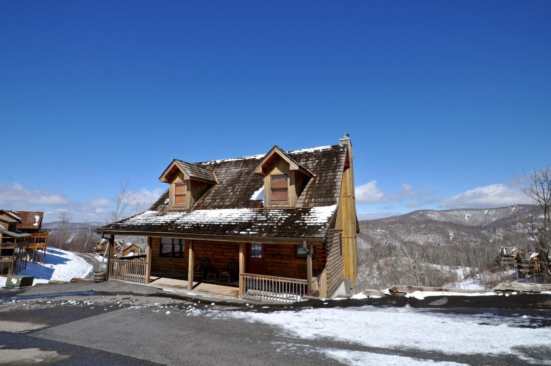 A wooden log cabin with a porch and a small attached structure, set against a mountainous backdrop.