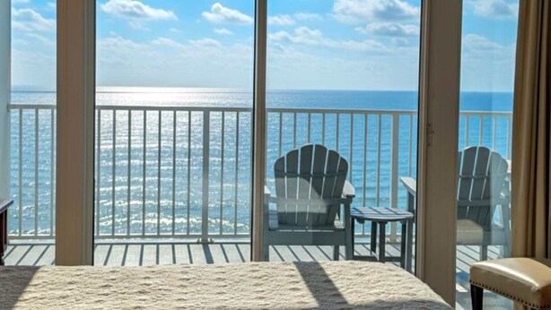 A room with a large window overlooking the ocean, a deck with chairs, and a small table. - Lighthouse 1405 (Gulf Shores)