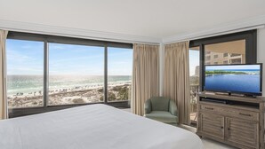 Habitación de hotel con una cama grande, televisión, aparador de madera y vista a la playa y al océano.