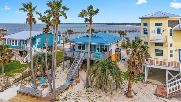 A beachfront property with multiple buildings, including a house with a blue roof and a yellow building with a balcony. - Tortuga Bay | 2BR/1BA Waterfront Home | Pet-friendly Fort Morgan Retreat (Gulf Shores)
