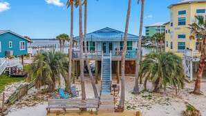 A beachfront property with a wooden deck, a staircase leading to a balcony, and palm trees. - Tortuga Bay | 2BR/1BA Waterfront Home | Pet-friendly Fort Morgan Retreat (Gulf Shores)