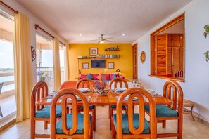 Een eetgedeelte met een houten tafel en stoelen, een plafondventilator en uitzicht op zee door grote ramen.