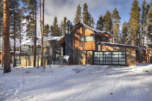 Una cabaña de troncos moderna con garaje, rodeada de nieve y altos pinos.