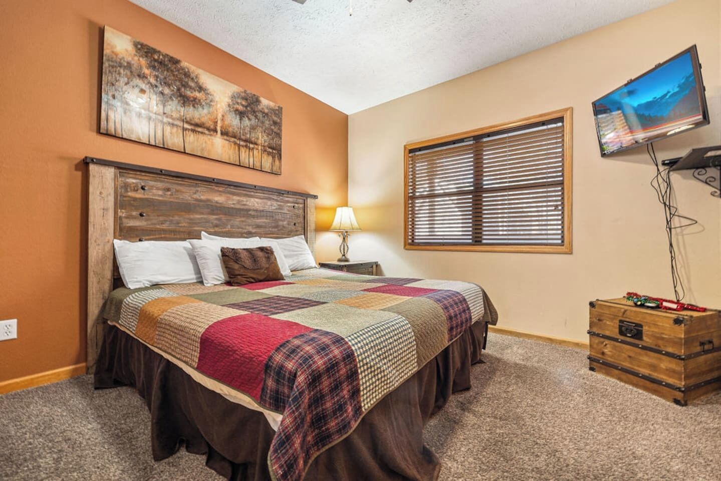 A bedroom with a wooden headboard, a quilted bedspread, a nightstand with a lamp, a window with blinds, and a television on the wall.