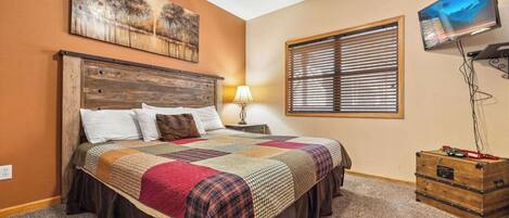 A bedroom with a wooden headboard, a quilted bedspread, a nightstand with a lamp, a window with blinds, and a television on the wall.