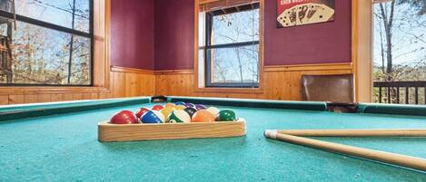 Une table de billard avec des billes et des queues, de grandes fenêtres donnant sur la forêt et un panneau accroché au mur.