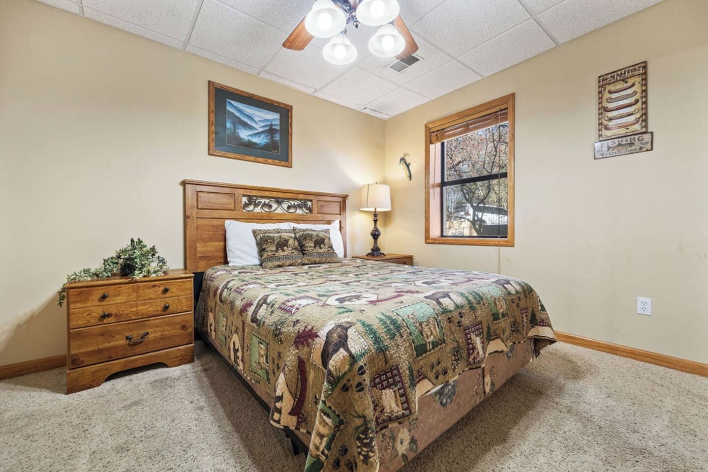 Un dormitorio con cabecera de madera, cama con colcha estampada, mesita de noche con lámpara, cómoda y ventana con vistas.