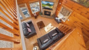 A log cabin interior with a stone fireplace, wooden walls, a large window, and a view of the outdoors.