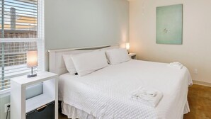 A bedroom with a bed, a ceiling fan, a bedside table with a lamp, and a window with blinds. - Updated Coastal Duplex | Heated Pool & Walk to the Beach (Corpus Christi)