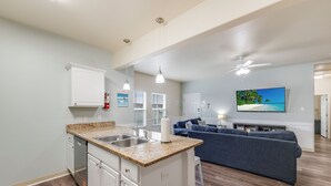 A modern kitchen with a granite countertop, white cabinets, and a dishwasher. There is a dining area with a blue sofa and a flat-screen TV mounted on the wall. - Updated Coastal Duplex | Heated Pool & Walk to the Beach (Corpus Christi)