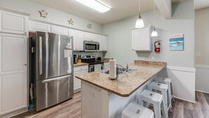 A modern kitchen with white cabinetry, stainless steel appliances, a granite countertop, and a small island with bar stools. - Updated Coastal Duplex | Heated Pool & Walk to the Beach (Corpus Christi)