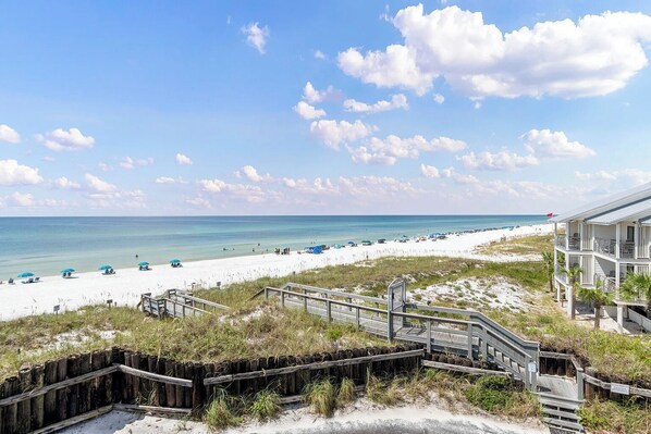 A beach view with a wooden boardwalk leading to a building with multiple balconies overlooking the ocean.