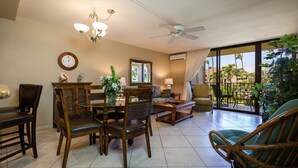 A spacious living area with a dining table, chairs, a sofa, and a wicker chair. There is a ceiling fan, a chandelier, and a large window with a view of palm trees.