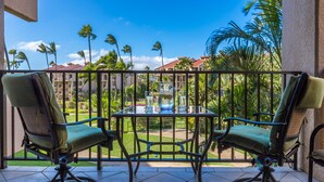 A balcony with two chairs and a table, overlooking a garden with palm trees and a building in the background.