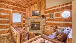 A log cabin interior with a stone fireplace, wooden furniture, and a television mounted above it.