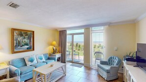 A living room with a sofa, coffee table, and armchairs. There is a painting on the wall, a lamp, and a TV on a cabinet. The room has a sliding glass door leading to a balcony with a view of the beach. - Villa 187 - Amore by the Sea 2 Bedroom, 1 Bathroom (Jekyll Island)