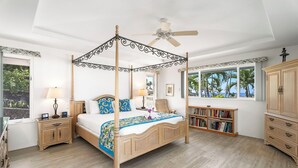 Uma cama com dossel, um ventilador de teto, uma estante de livros e vista para palmeiras através de janelas amplas.