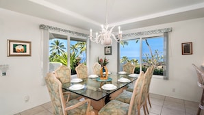 Uma sala de jantar com mesa de vidro, cadeiras com padrão tropical, um candelabro e grandes janelas com vista para palmeiras e o oceano.