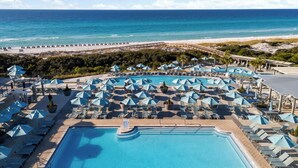 A resort pool area with multiple sun loungers and umbrellas, a large central pool, and a beach view.