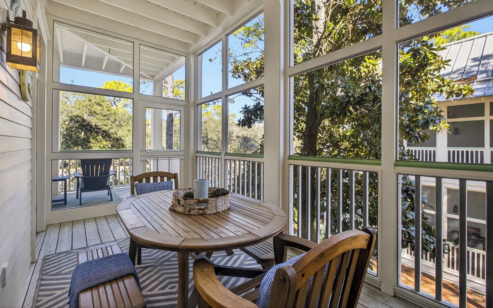 A covered outdoor patio with a wooden table and chairs, surrounded by large windows offering views of trees and a house.