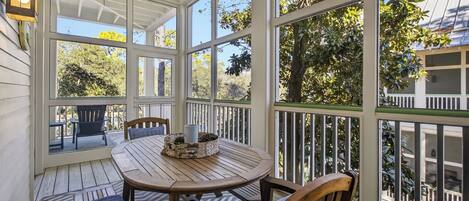 A covered outdoor patio with a wooden table and chairs, surrounded by large windows offering views of trees and a house.