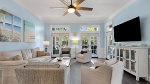 A spacious living room with a ceiling fan, a large TV, and a view through glass doors to the outdoors.