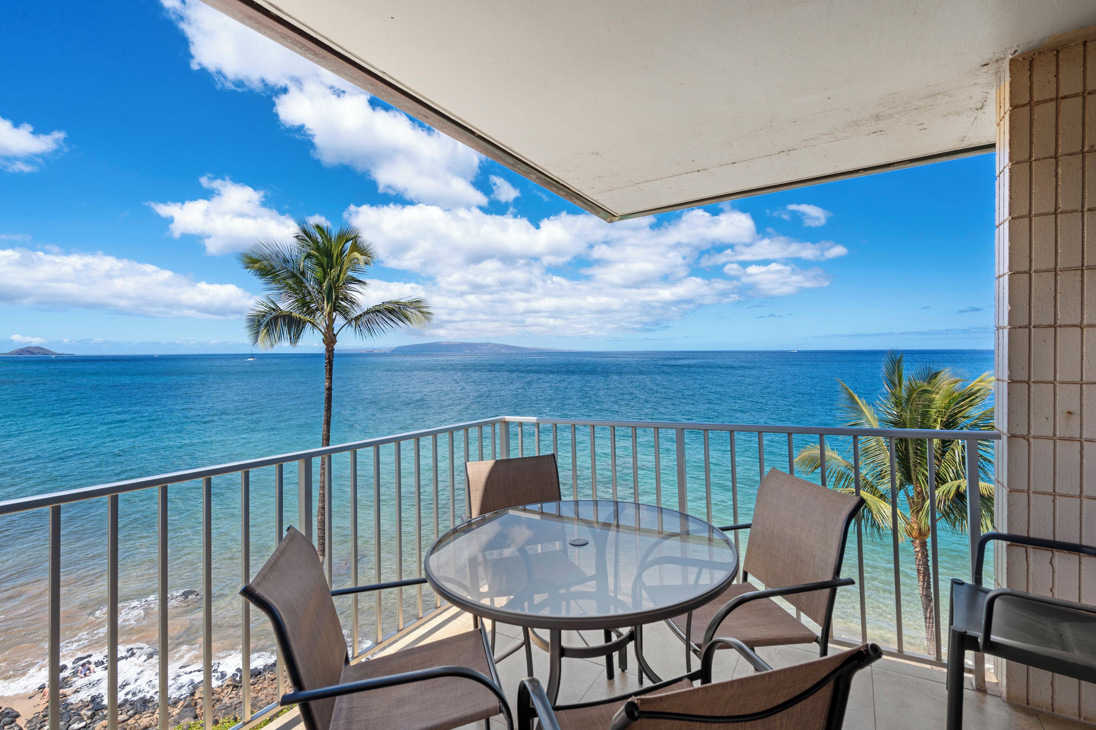 A balcony with a glass table and chairs overlooking a beach with clear blue water and a palm tree.