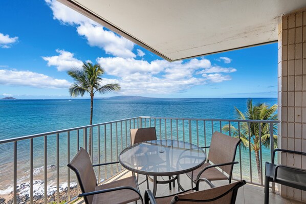 A balcony with a glass table and chairs overlooking a beach with clear blue water and a palm tree.