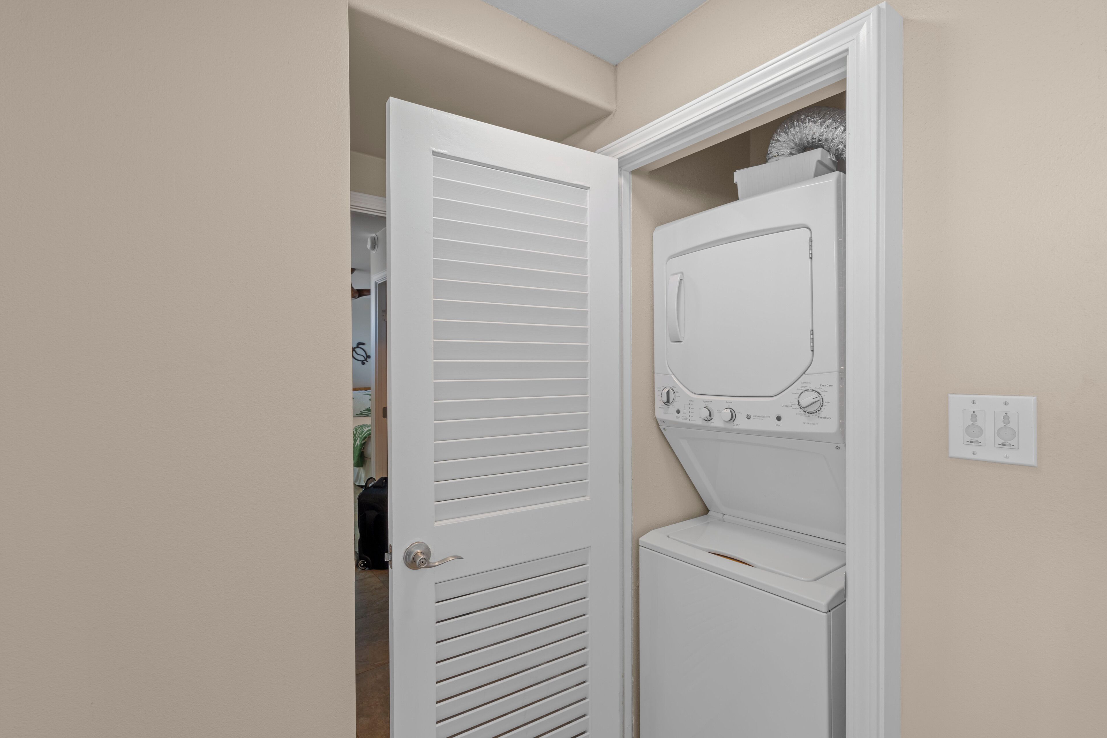 A laundry room with a front-loading washer and dryer, a door with horizontal slats, and a wall-mounted electrical outlet.