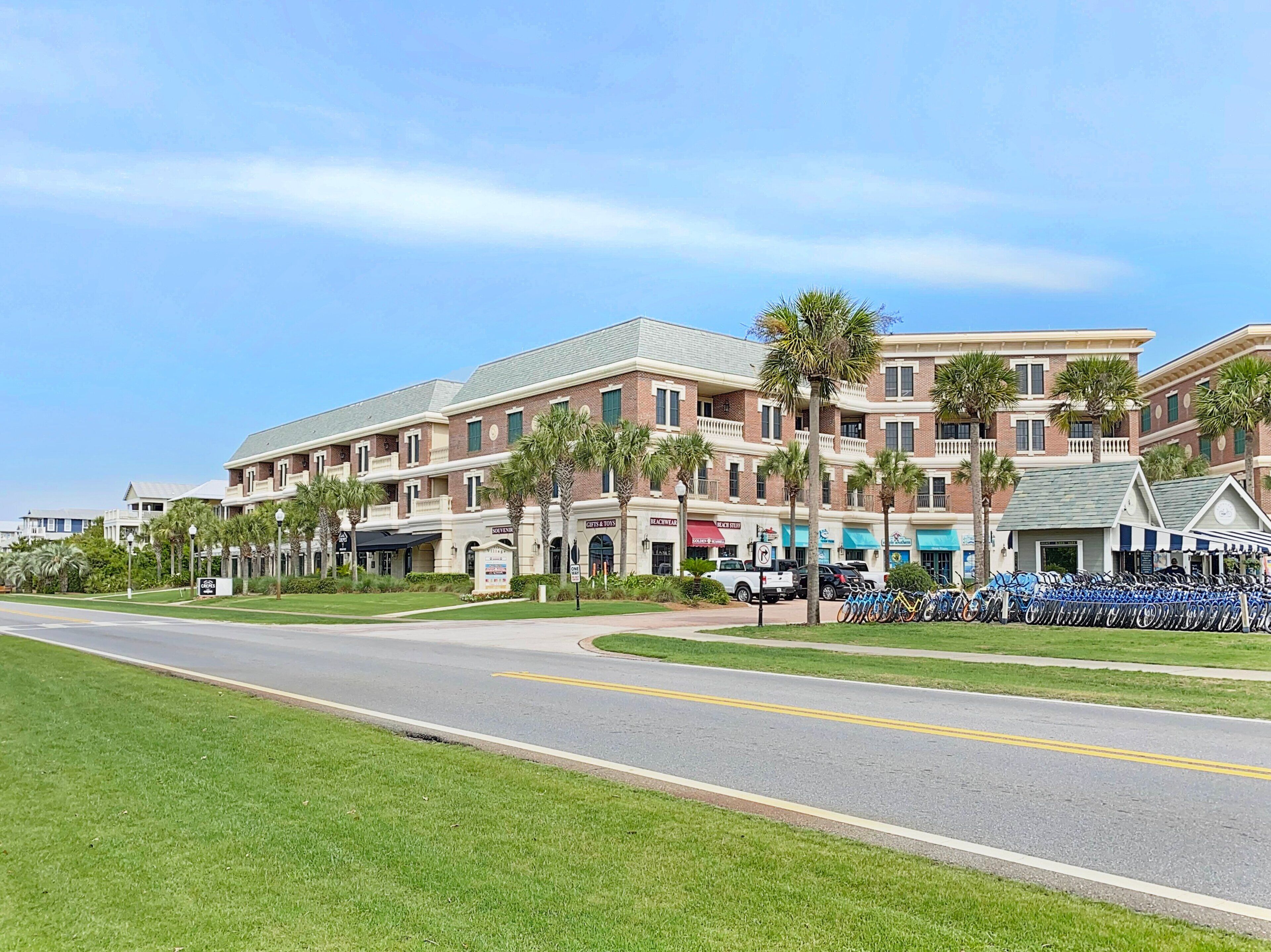 A multi-story hotel with a brick facade, palm trees, and a row of shops on the ground floor.
