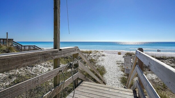 Un paseo marítimo de madera que lleva a una playa con cielos despejados y el océano al fondo.