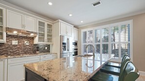 Una cocina moderna con armarios blancos, encimera de granito, electrodomésticos de acero inoxidable y un amplio ventanal con persianas.