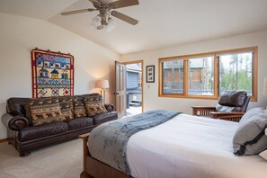 Un dormitorio con cama, sofá de cuero, colcha, ventilador de techo y ventana con vista a una cabaña.