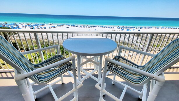 A balcony with two striped chairs and a small table, overlooking a beach with numerous umbrellas and people.