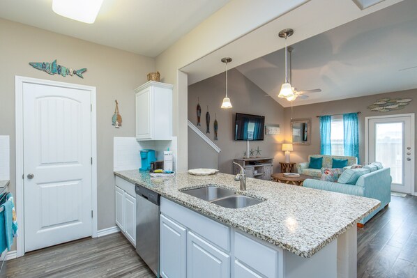 A kitchen with granite countertops, white cabinets, and a dishwasher. There is a dining area with a blue sofa and a television. - Family Getaway | Heated Pool, Patio & Play Area (Corpus Christi)