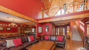 A spacious living area with a high ceiling, wooden beams, a large brown leather sofa set, a wooden coffee table, and a staircase leading to an upper level. - Spacious Mountain Escape w/ Hot Tub and Fireplace (OAKLAND)