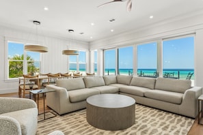 A spacious living room with a large sectional sofa, a round coffee table, and a dining area with wooden chairs and a table. Large windows offer a view of the ocean.