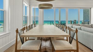 Een ruime eethoek met een lange houten tafel en stoelen, grote ramen met uitzicht op de oceaan en een moderne hanglamp.