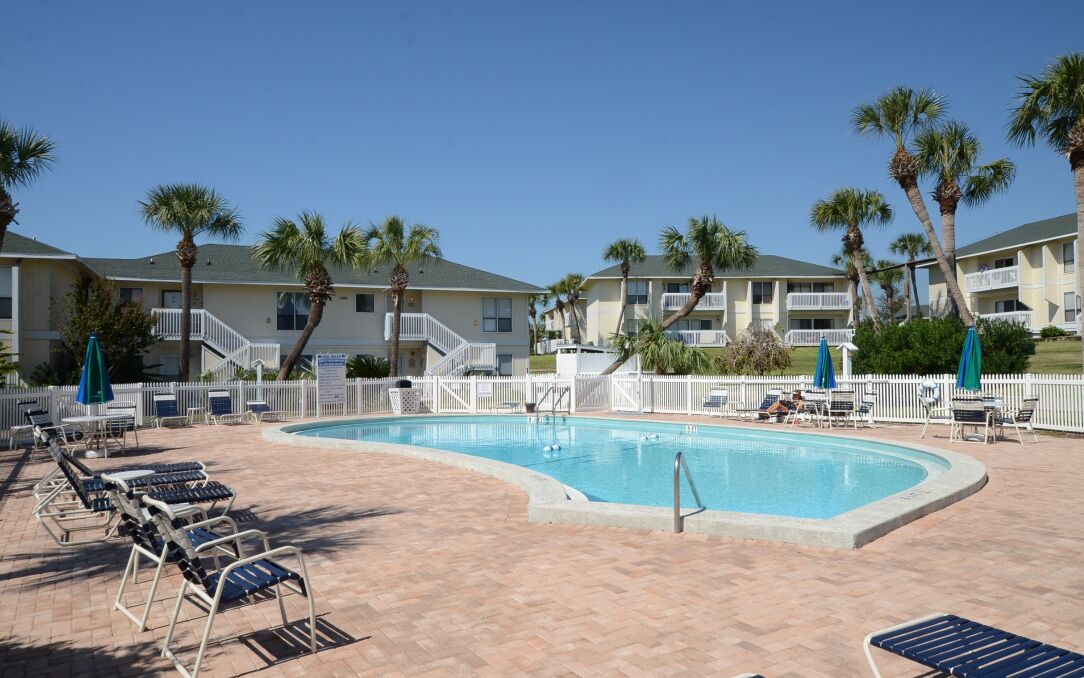 A swimming pool with lounge chairs and palm trees, surrounded by a white fence and buildings with balconies.