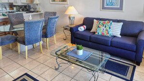 A living room with a blue sofa, matching chairs, a glass coffee table with a plant, a dining area with a table and chairs, and a kitchen in the background. - Budget Friendly 2Bd Beachfront with $1000000 View! Seasonal Beach Service! (Fort Walton Beach)