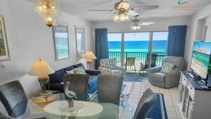 A spacious living room with a glass table, comfortable seating, a TV, and a balcony with a view of the ocean. - Budget Friendly 2Bd Beachfront with $1000000 View! Seasonal Beach Service! (Fort Walton Beach)