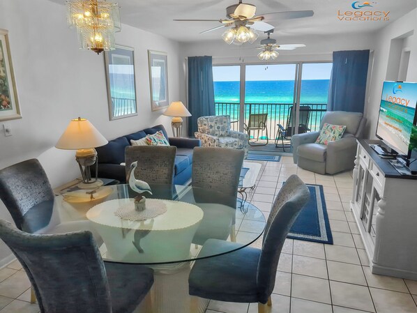 A spacious living room with a glass table, comfortable seating, a TV, and a balcony with a view of the ocean.