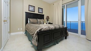 A bedroom with a large bed, a nightstand with a lamp, and a view of the ocean through a sliding glass door.