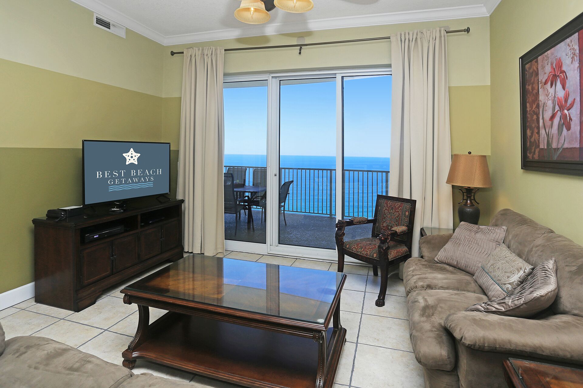 Una sala de estar con una televisión mostrando 'BEST BEACH GETAWAYS', un sofá, una mesa de centro, una silla y un balcón con vistas al océano.