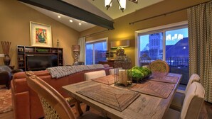 Ein gemütliches Wohnzimmer mit einer Couch, einem Esstisch mit Platzsets, einem Fernseher und einem großen Fenster mit Blick auf ein Gebäude.