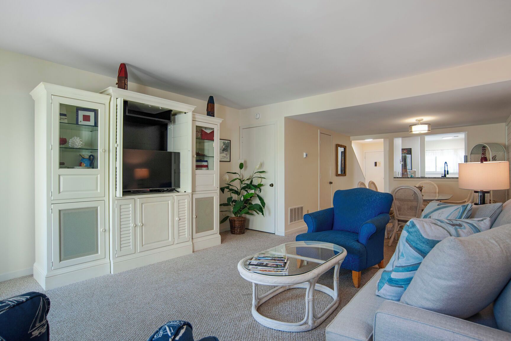 A living room with a television, a blue armchair, a grey sofa, a glass coffee table, and a white cabinet with glass doors.