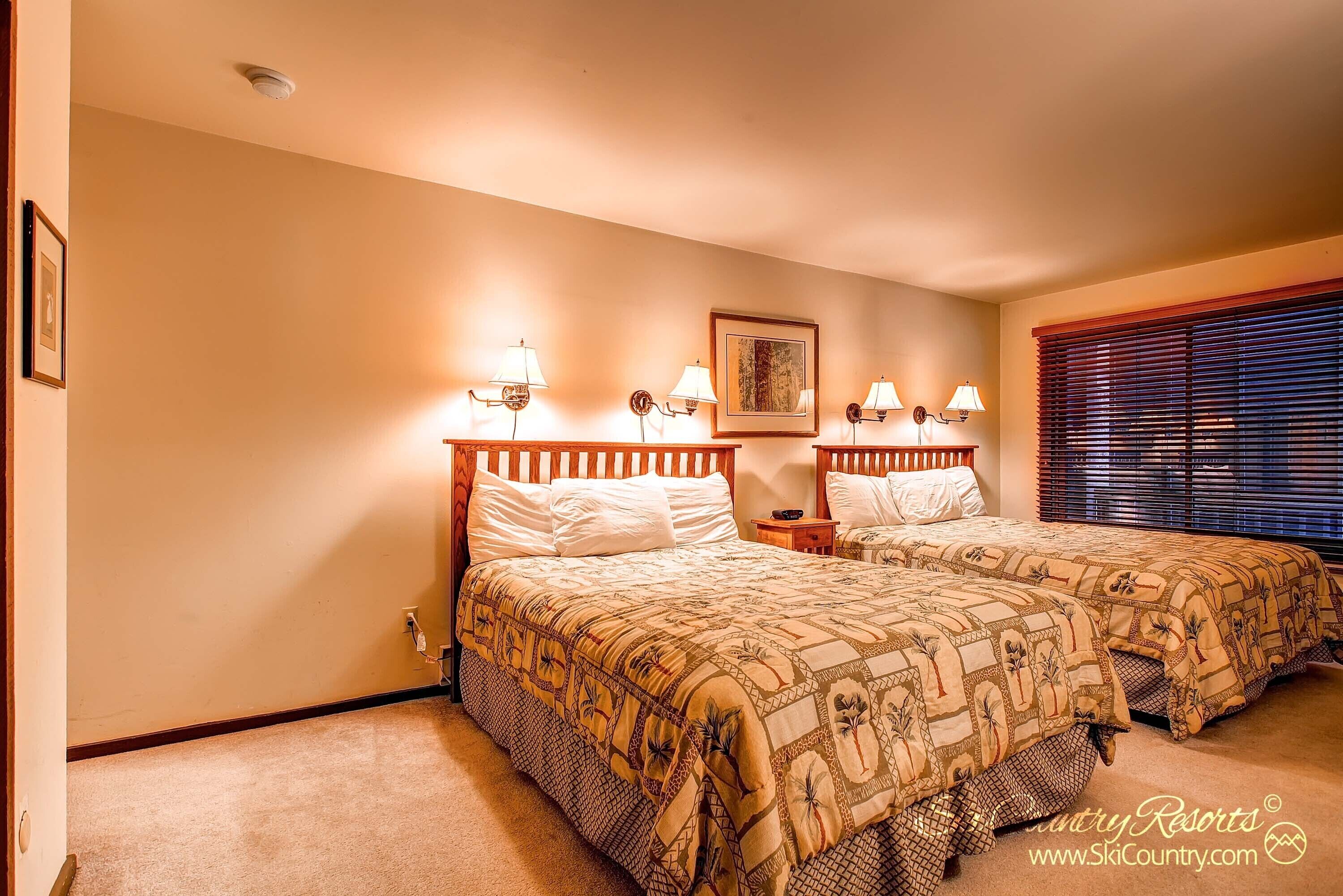 A bedroom with two beds, each with a patterned bedspread, bedside lamps, a framed picture on the wall, and a window with blinds.