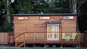 A wooden cabin with a deck, two windows, and a small porch area with chairs. - Modern Cozy Urban Tiny House near Airport and Transportation-Private Setting (Des Moines)