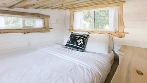 A bedroom with a wooden ceiling, a bed with white bedding, a window with curtains, and a wooden nightstand. - Modern Cozy Urban Tiny House near Airport and Transportation-Private Setting (Des Moines)